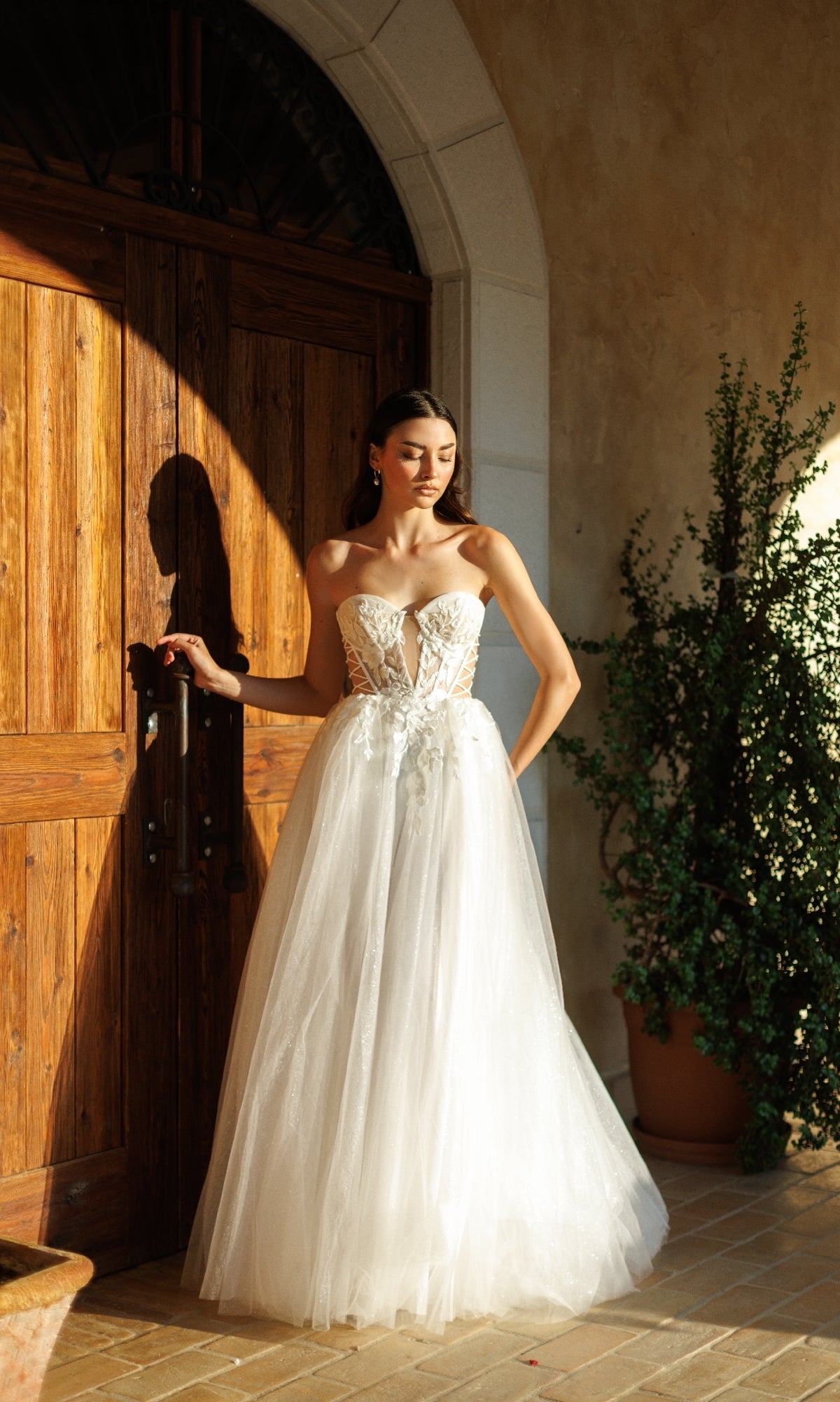 A woman in the Tina Holly TW480 long formal dress with a tulle A-line skirt and corset bodice stands by a large wooden door, sunlight casting shadows while a potted plant sits nearby.